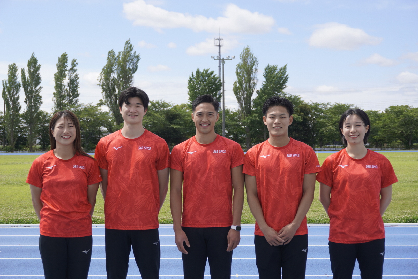 オレンジのユニフォームを着た5人の選手が青いトラックの上に並んで立ち背景に芝生と並木が見える