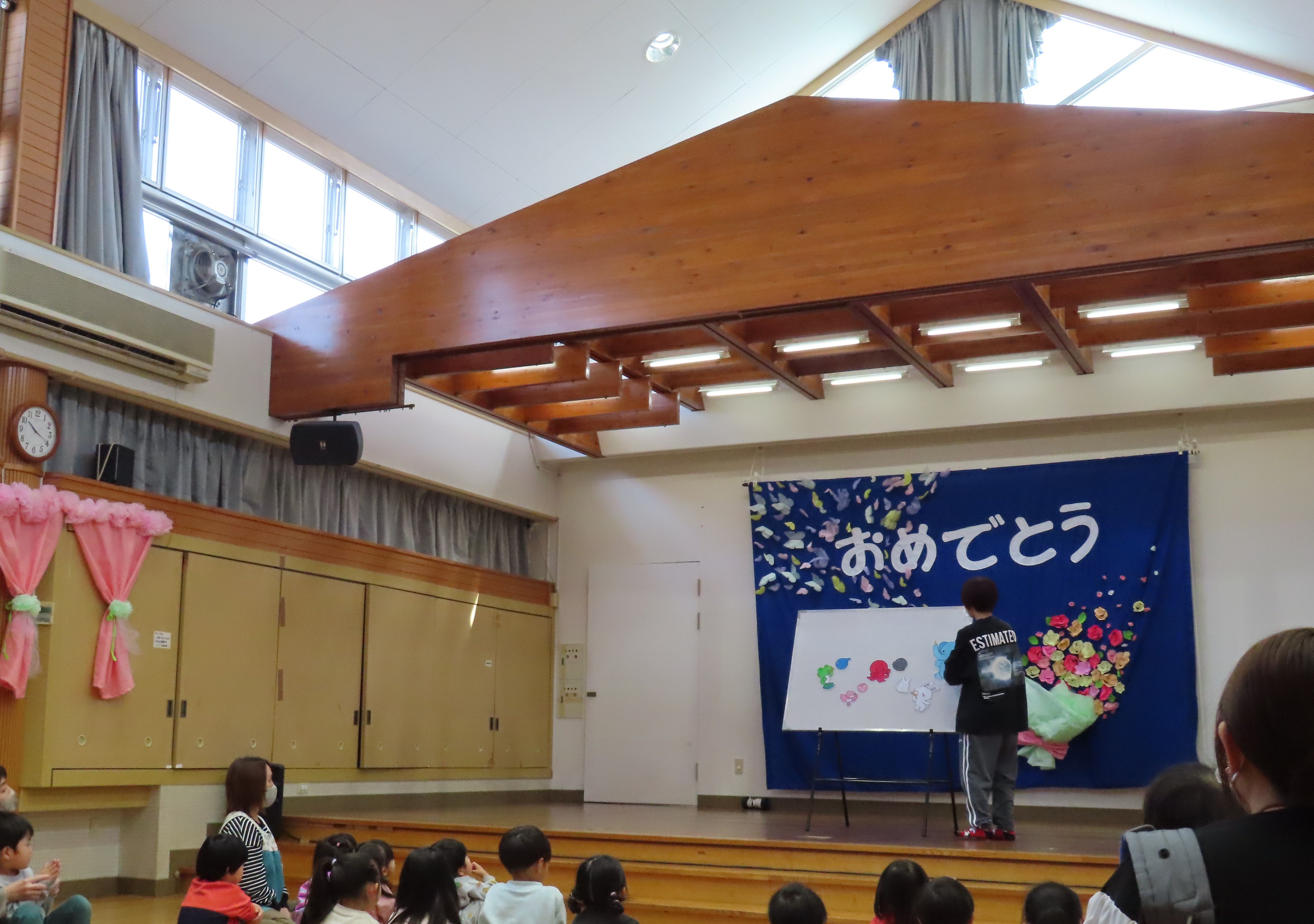 こどもたちが集まる教室で、黒い服を着た人物がホワイトボードに絵を描いている様子を示す画像。