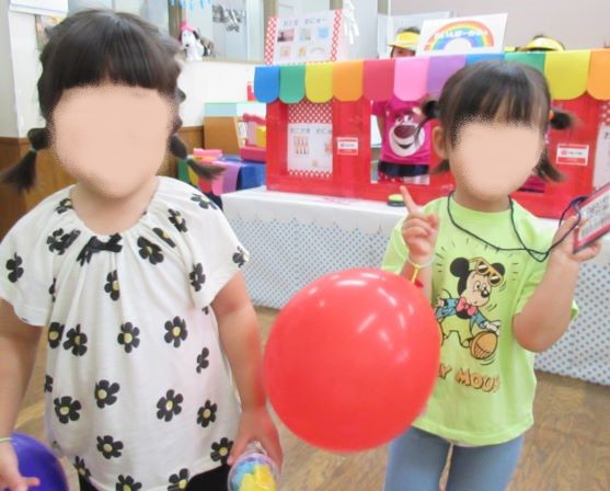 二人のこどもが赤い風船を持ち、カラフルな背景の中で遊んでいる様子。