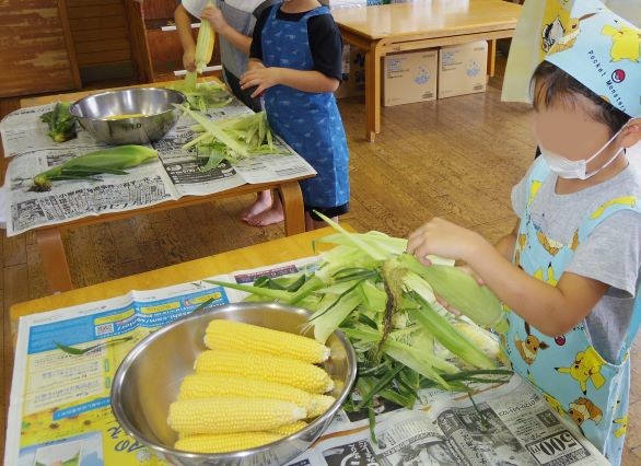 こどもがテーブルでトウモロコシの皮を剥いている様子が写っている画像。