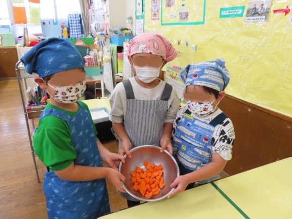 三人のこどもたちがオレンジ色の人参を入れたボウルを持っている教室の様子。