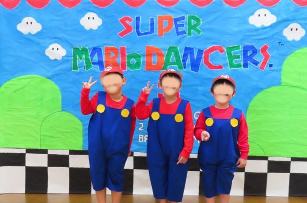 スーパーマリオの衣装を着た3人のこどもたちがポーズをとっている背景にはカラフルなバナーがある。