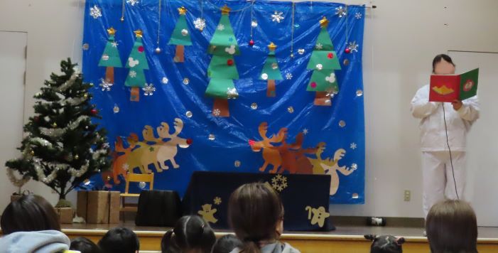 クリスマスの装飾が施された背景の前で、何かを持った人物が話している様子と、それを見ているこどもたち。
