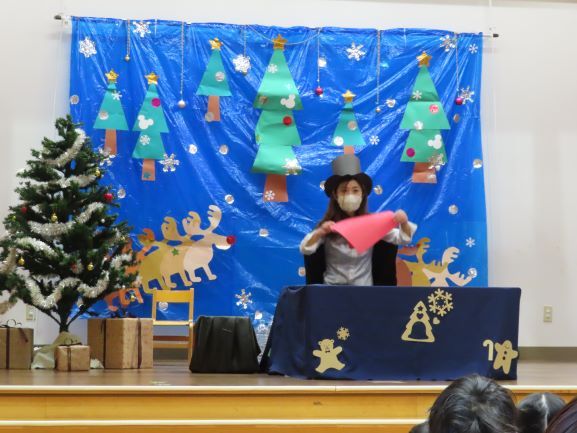 舞台で赤い布を持つマジシャンがクリスマスの装飾が施された背景の前でパフォーマンスをしている。