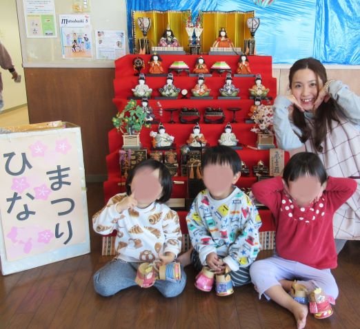 ひな祭りの飾りの前でポーズをとるこどもたちと大人の女性。