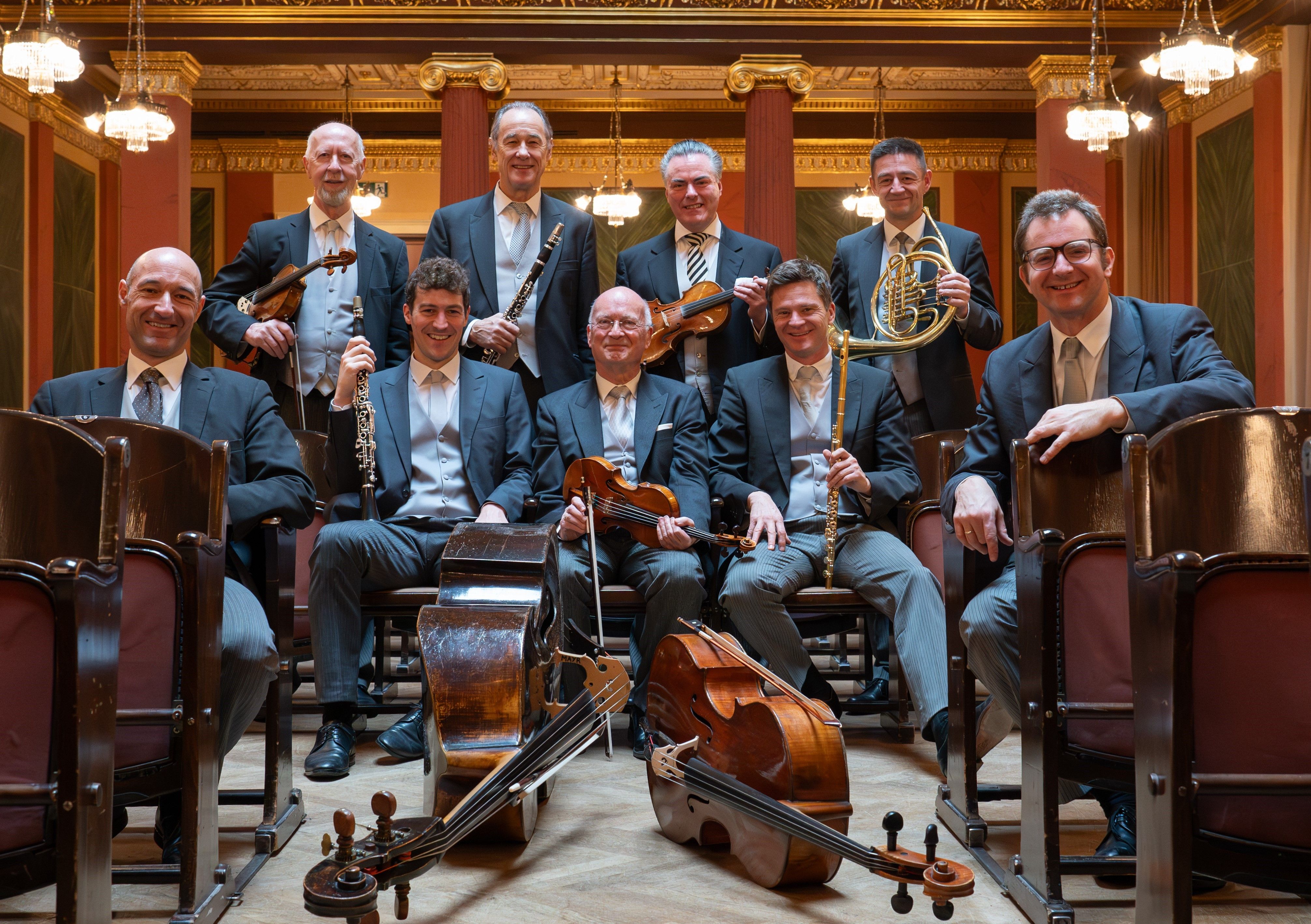 オーケストラの演奏者たちが楽器を持って座っている写真で、背景には豪華な内装が見える。