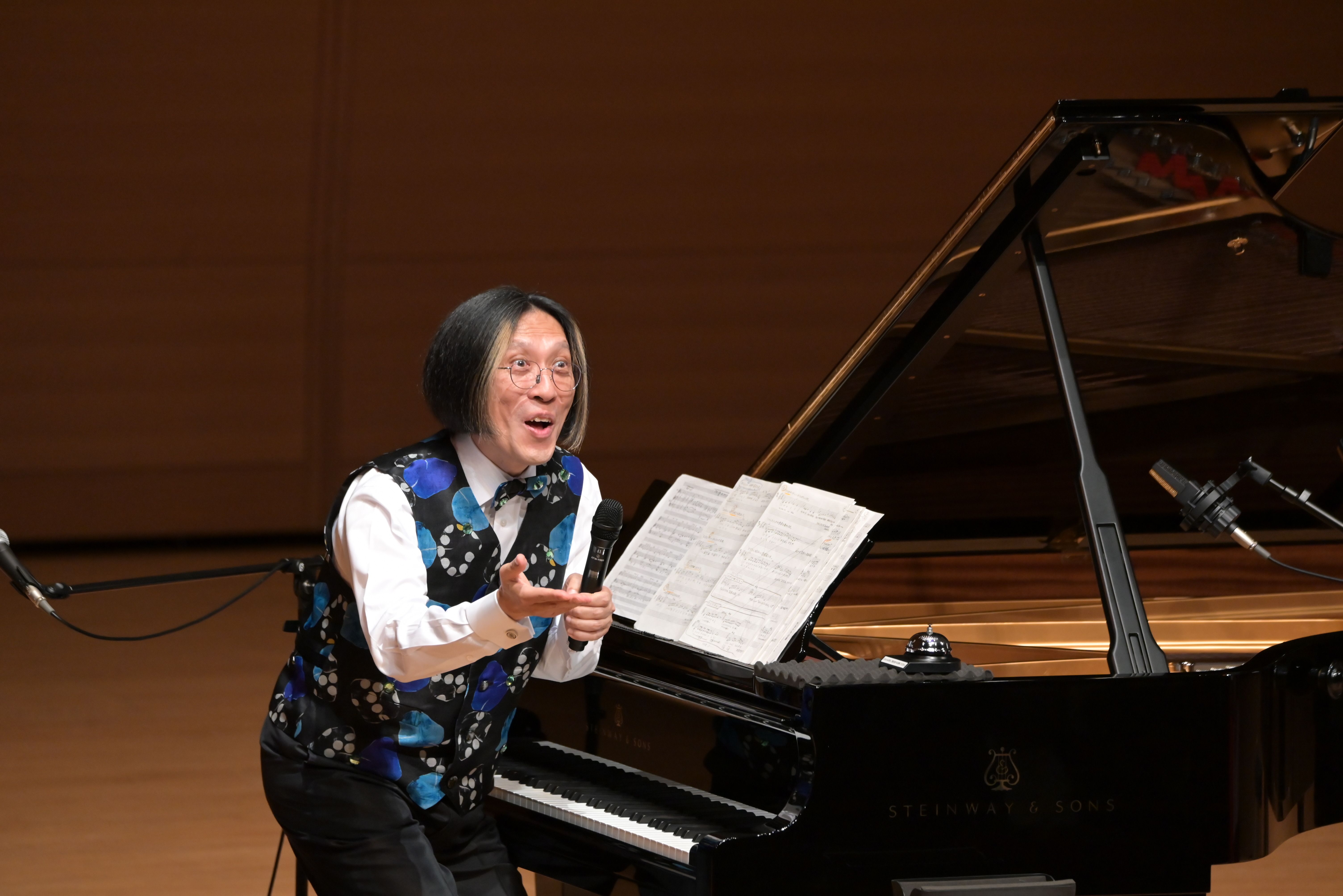 ピアノの前でマイクを持ちながら話している人物が写っている画像