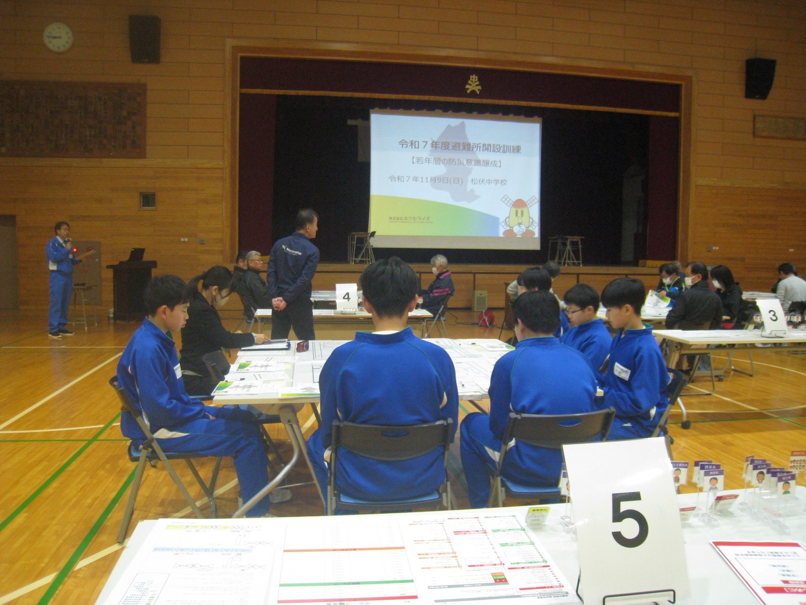 体育館で青い制服を着た生徒たちがテーブルを囲んでいる様子が映っている画像