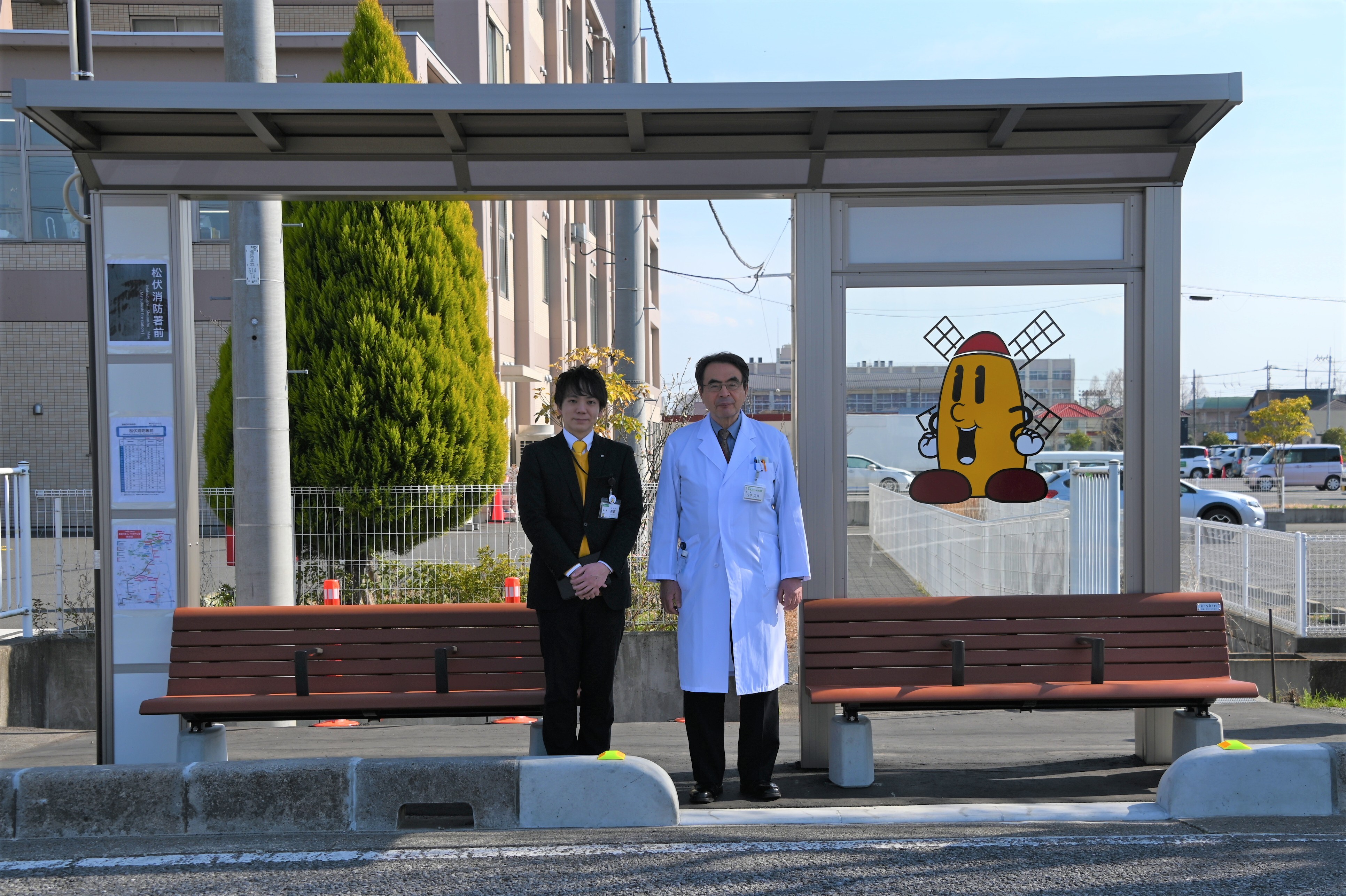 屋根付きのバス停のベンチにスーツ姿の人物と白衣の人物が並んで立ちガラスに風車のキャラクターのステッカーが貼られている背景に建物や駐車場が見える