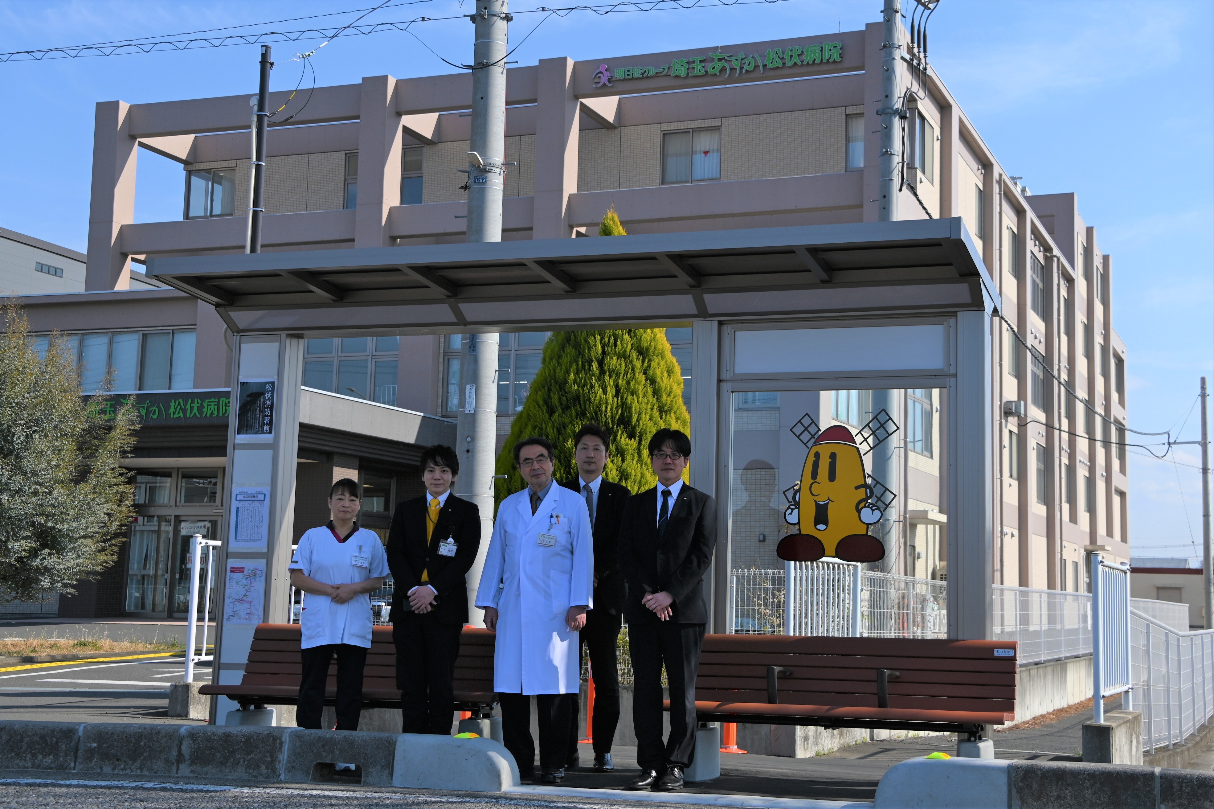 バス停の屋根の下に医療服や白衣とスーツを着た数人が並んで立ち背景にビルと風車をあしらったキャラクターのイラストが見える