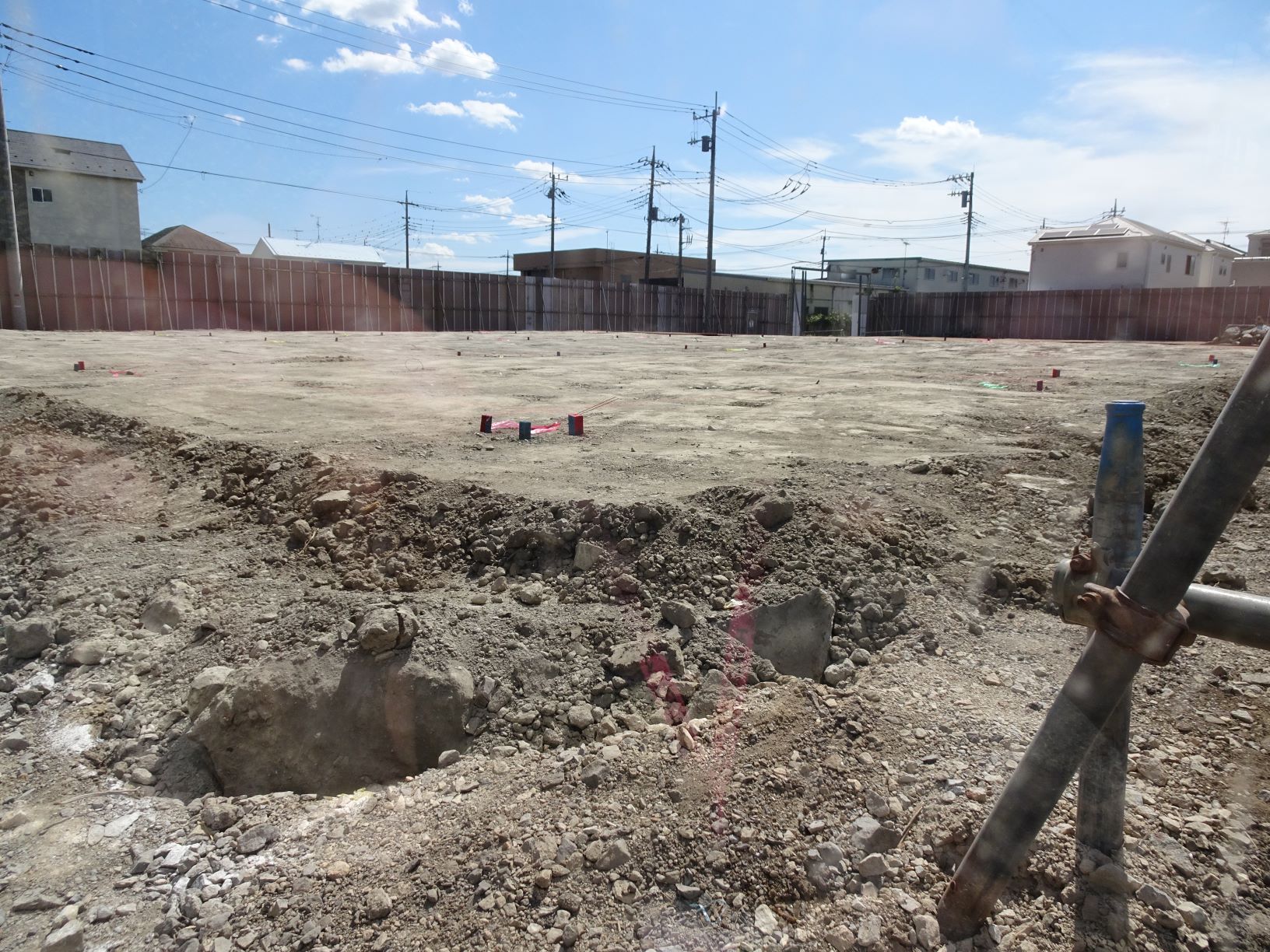 広い更地の工事現場の手前に赤いプラスチック片などが散らばる土砂の穴と砂利越しに遠くに低い建物と電柱が並ぶ青空の風景