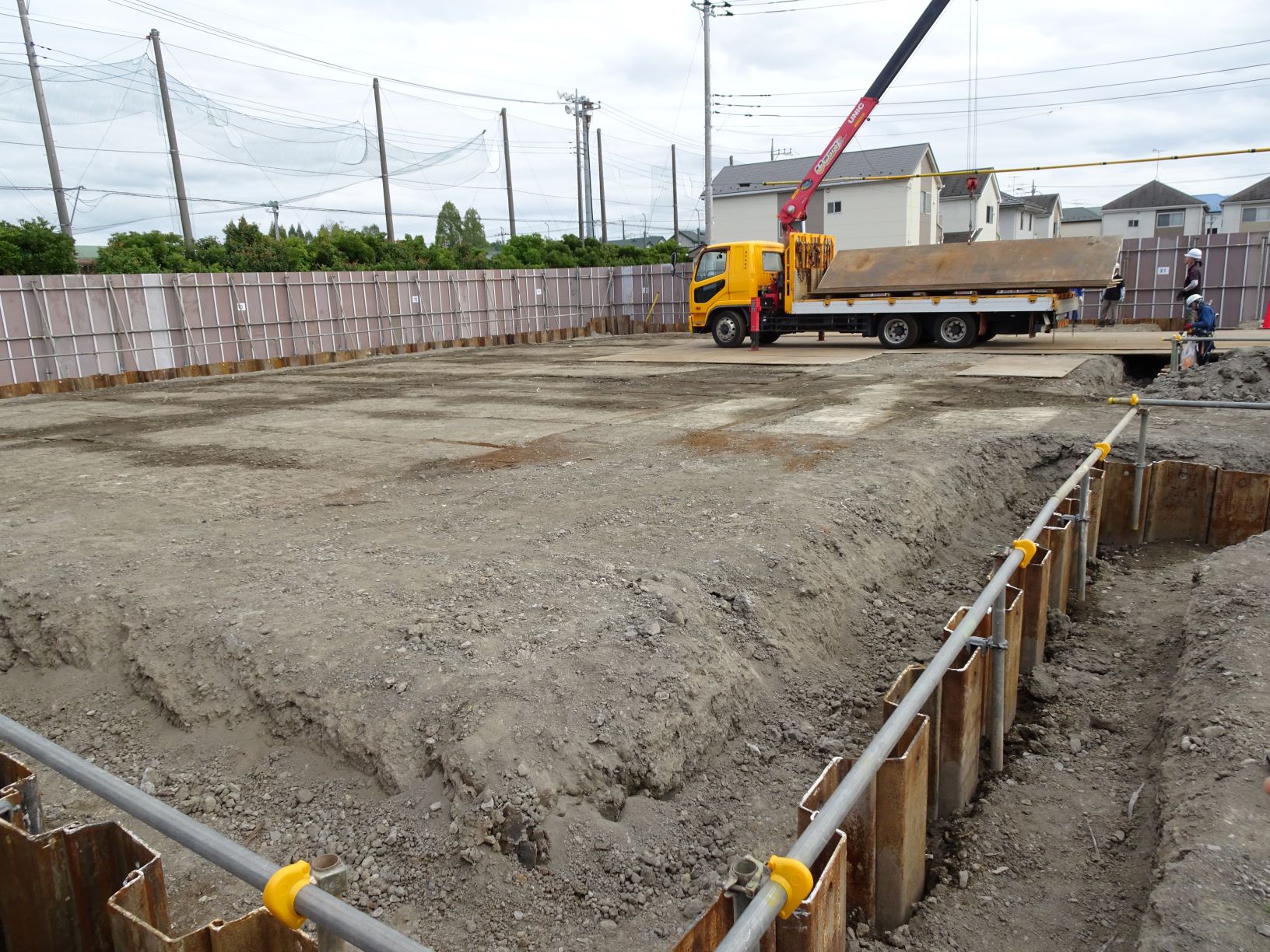 フェンスに囲まれた更地の建設現場の手前に仮設の型枠が並び奥にクレーン付きトラックが停車している風景