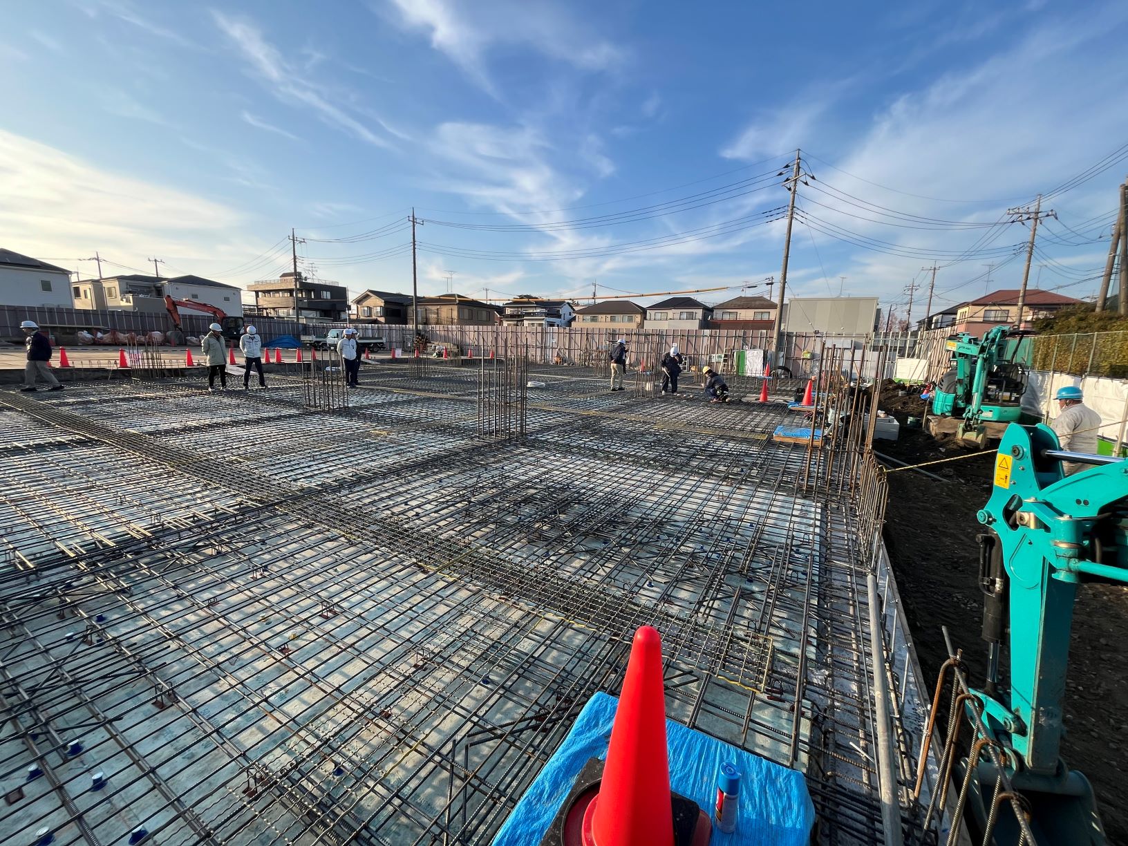 住宅地の現場で鉄筋が格子状に敷かれた基礎工事現場と作業員や重機が見える風景