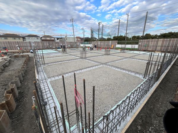 基礎工事中の建設現場で格子状に配された鉄筋と砕石の床面を作業者が点検している住宅地の風景