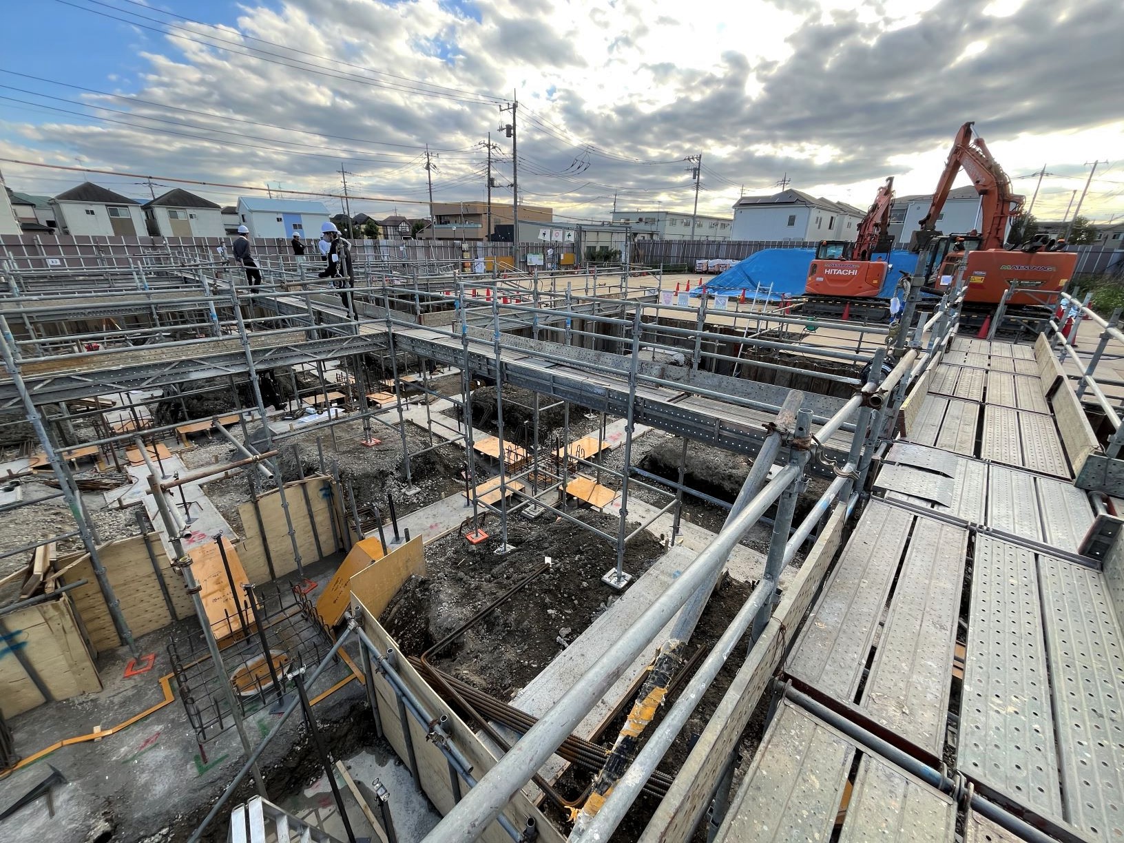 足場の上から見下ろす住宅建設現場と作業員がいる重機と仮設通路と雲のかかった空