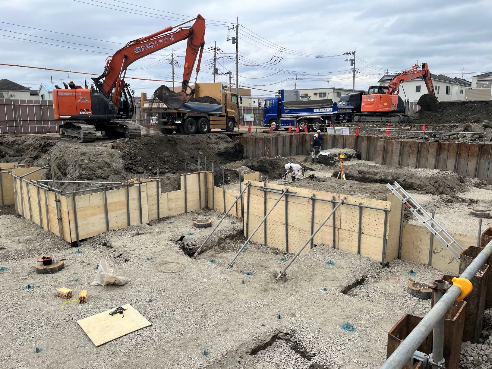 住宅地の工事現場で複数の重機と作業員が基礎工事を行っている様子