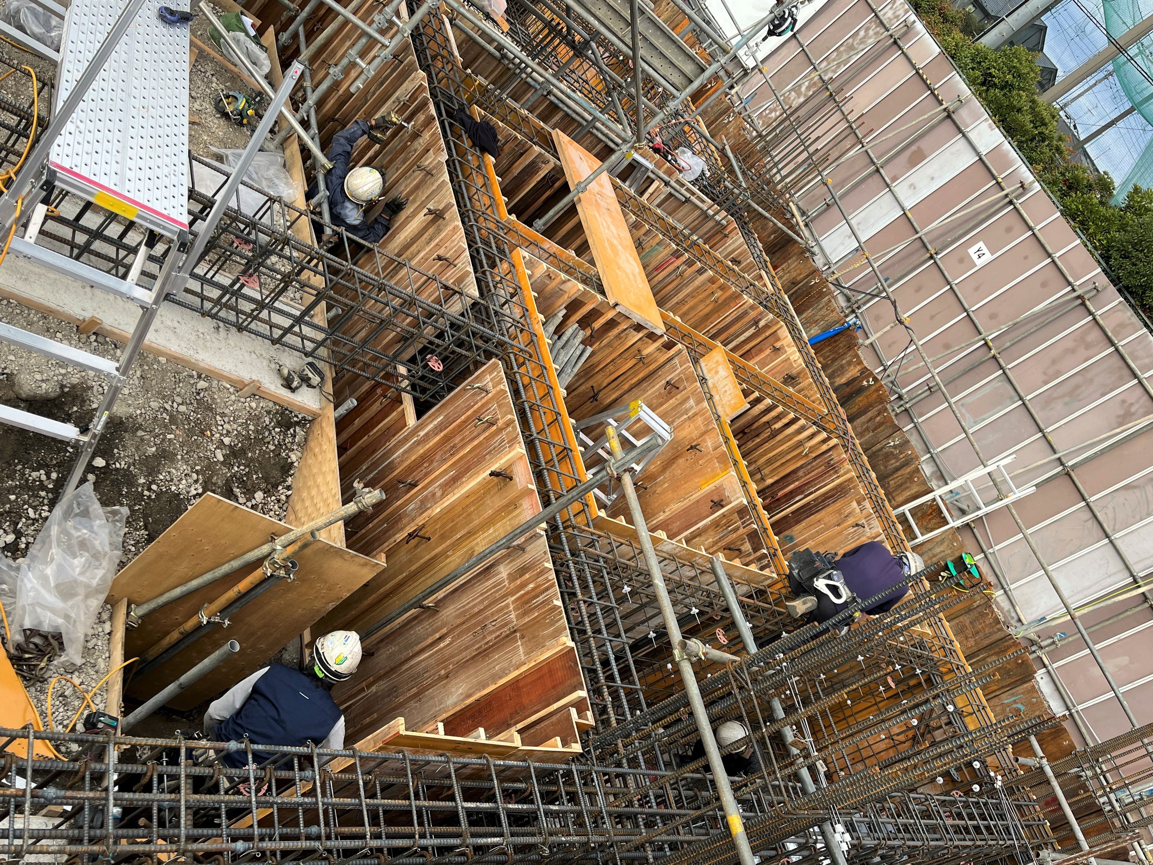 高層の足場と木製型枠の周りでヘルメットを着用した作業員が作業している建設現場の俯瞰写真