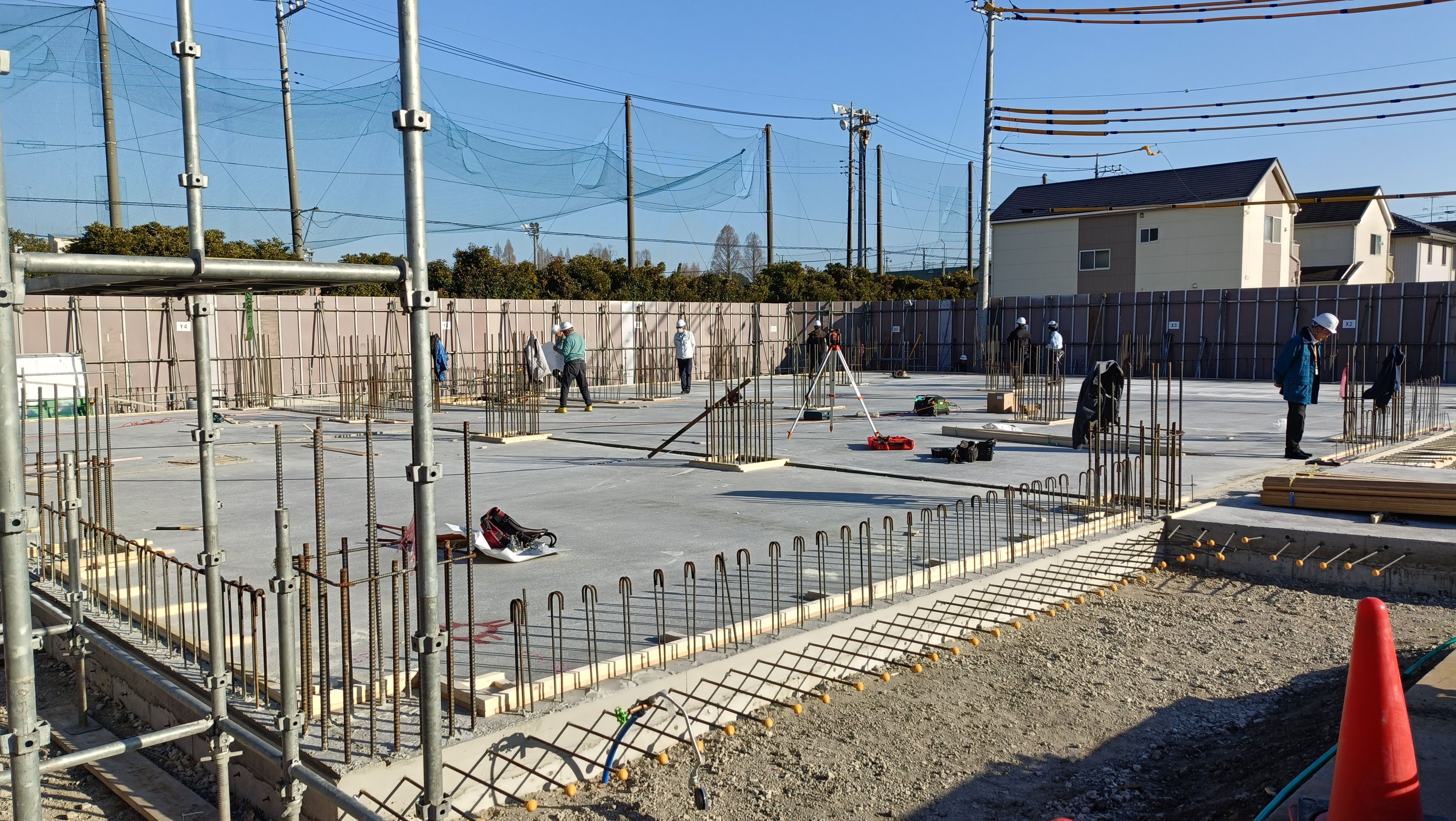 工事現場でコンクリート基礎の配筋と三脚などの測量機器が置かれ作業者がヘルメットを着用して広い基礎面で作業している様子