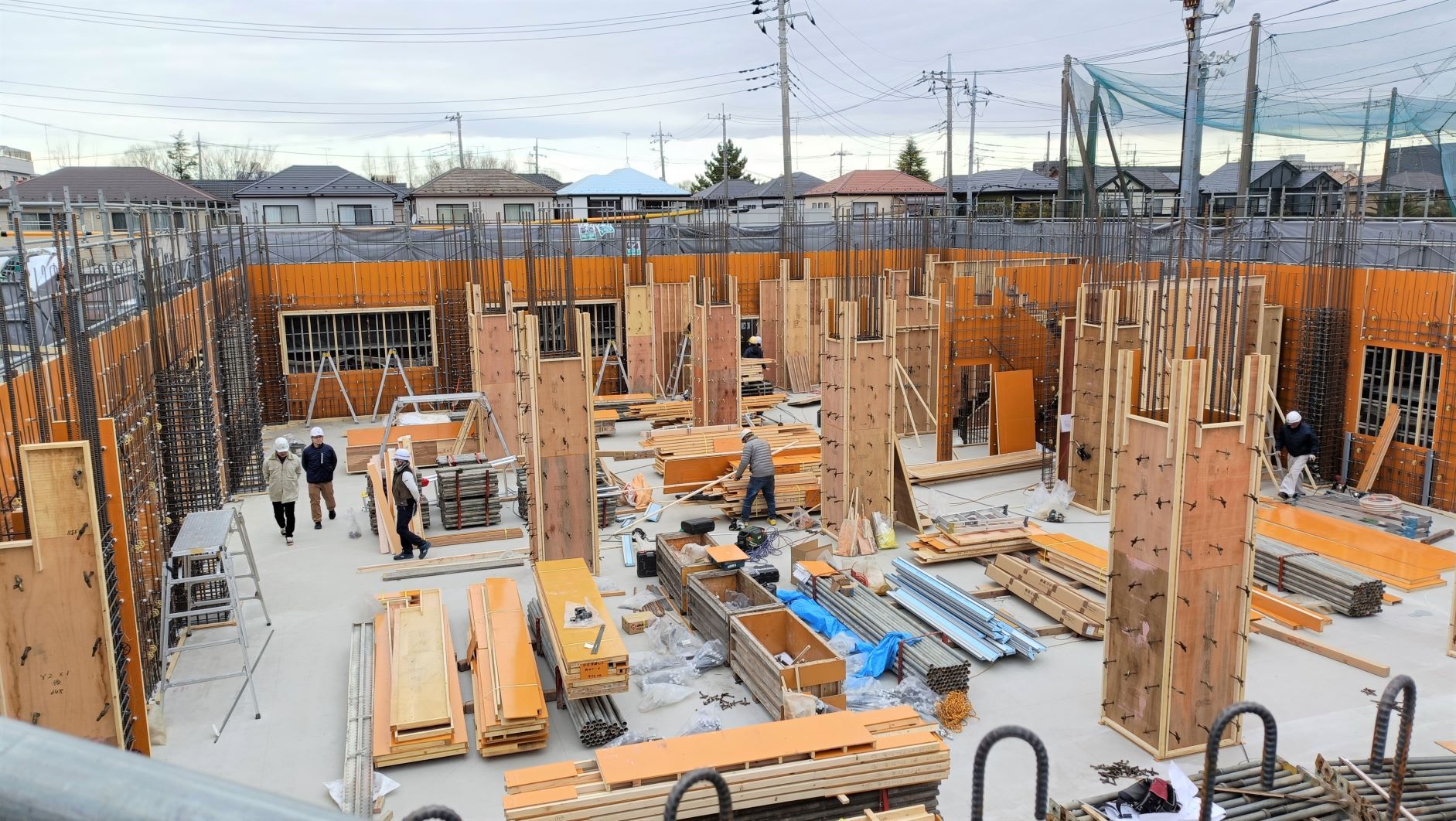 木造の柱と資材が並ぶ屋外の建設現場で数人の作業者が作業している様子