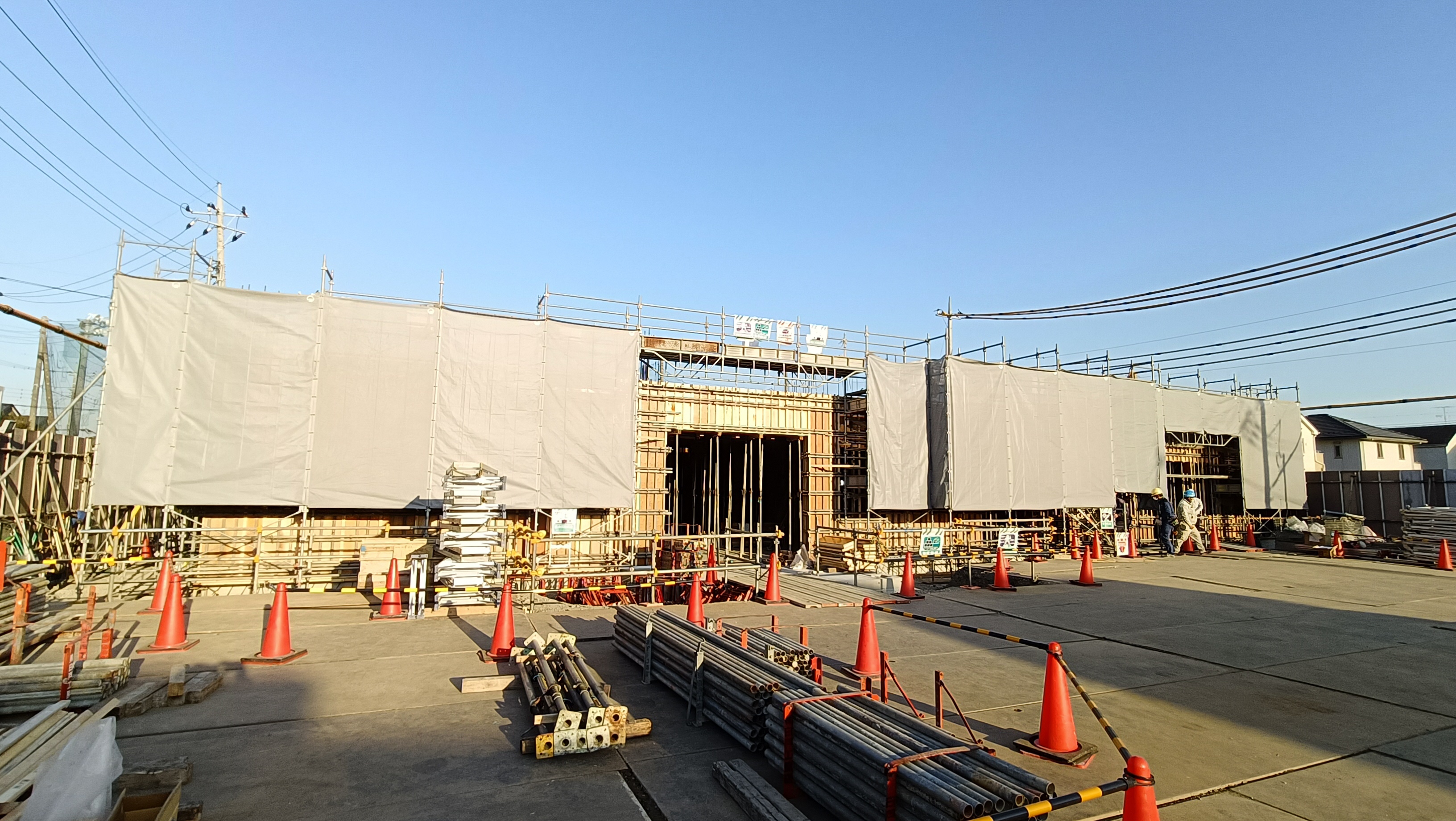 工事中の建物を覆う白いシートと足場の前に資材やオレンジのカラーコーンが並ぶ屋外の建設現場を広角で捉えた写真