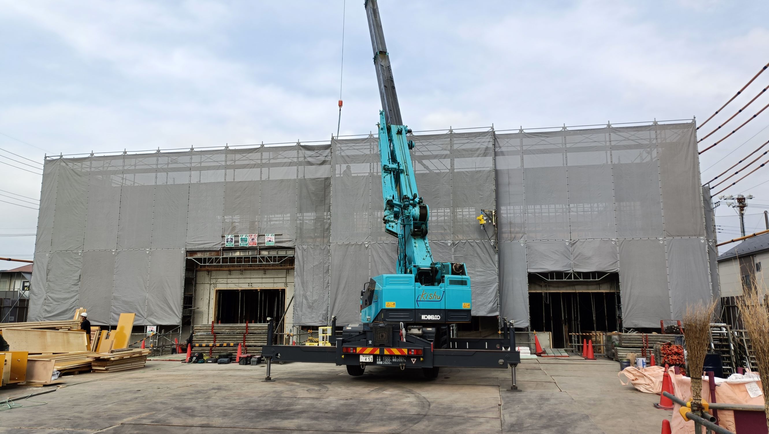 前面に青い大型クレーンが据えられた覆いのかかった建物の工事現場で作業員が足場や資材のそばで作業している全景