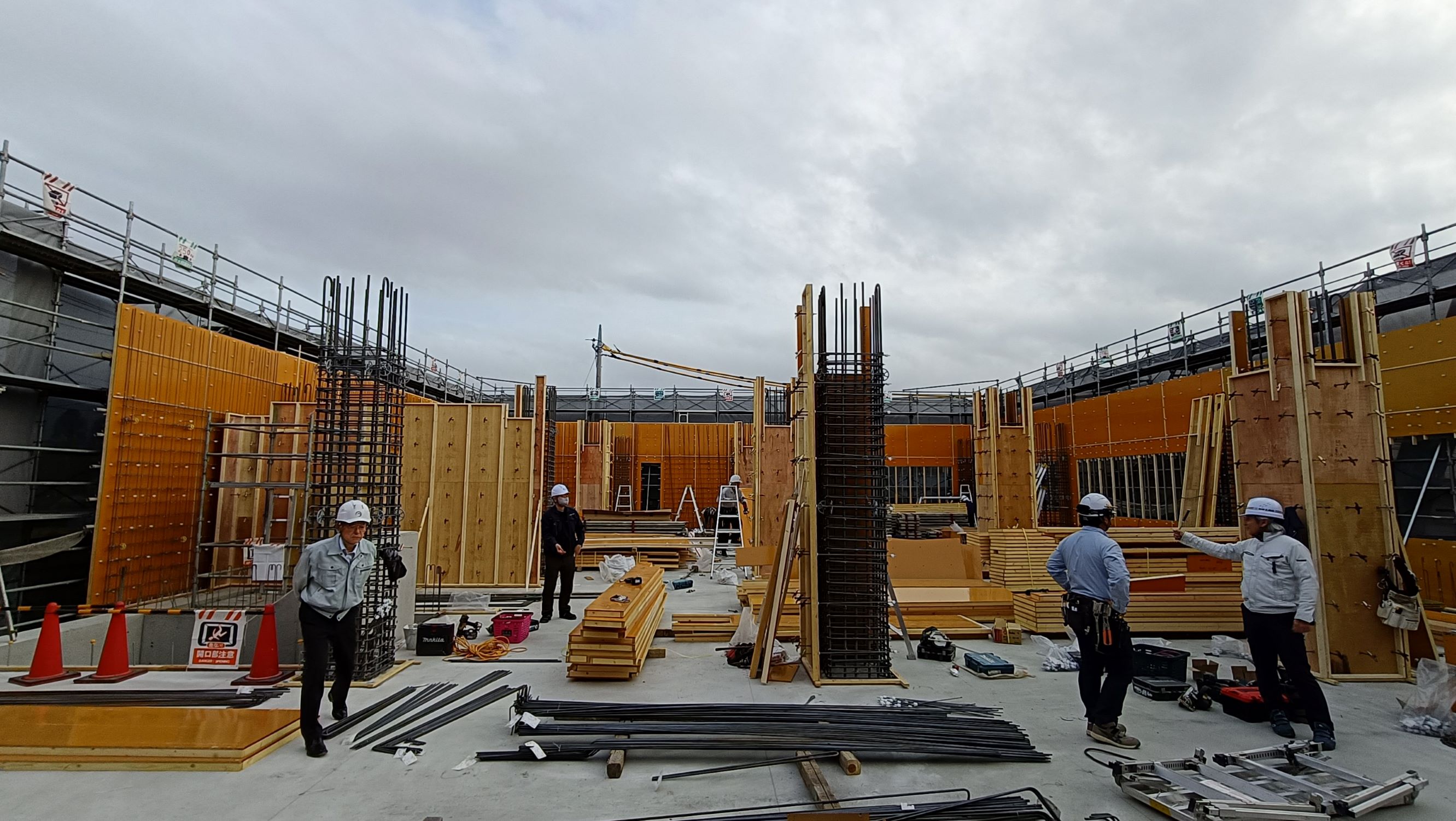 建設現場でヘルメットをかぶった作業員が木製の型枠や鉄筋資材に囲まれ作業や打ち合わせをしている広い作業エリアの風景