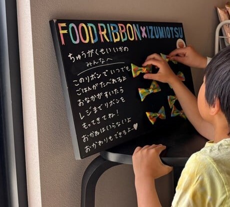 店内で黒いボードに掲示されたフードリボンがこどもによって手に取られている様子